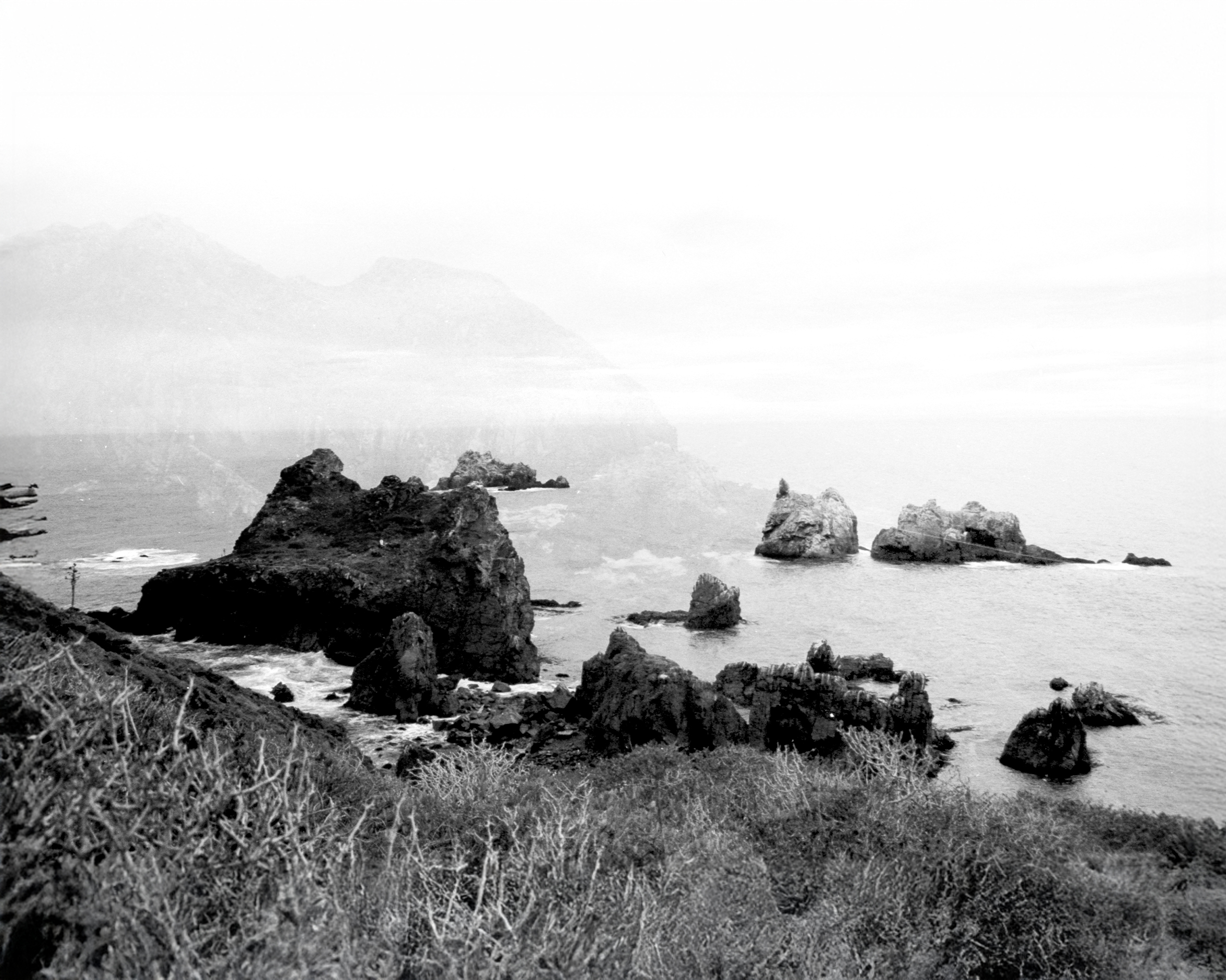 Fotografía en blanco y negro de costa rocosa con grandes rocas en el agua y montañas al fondo.
