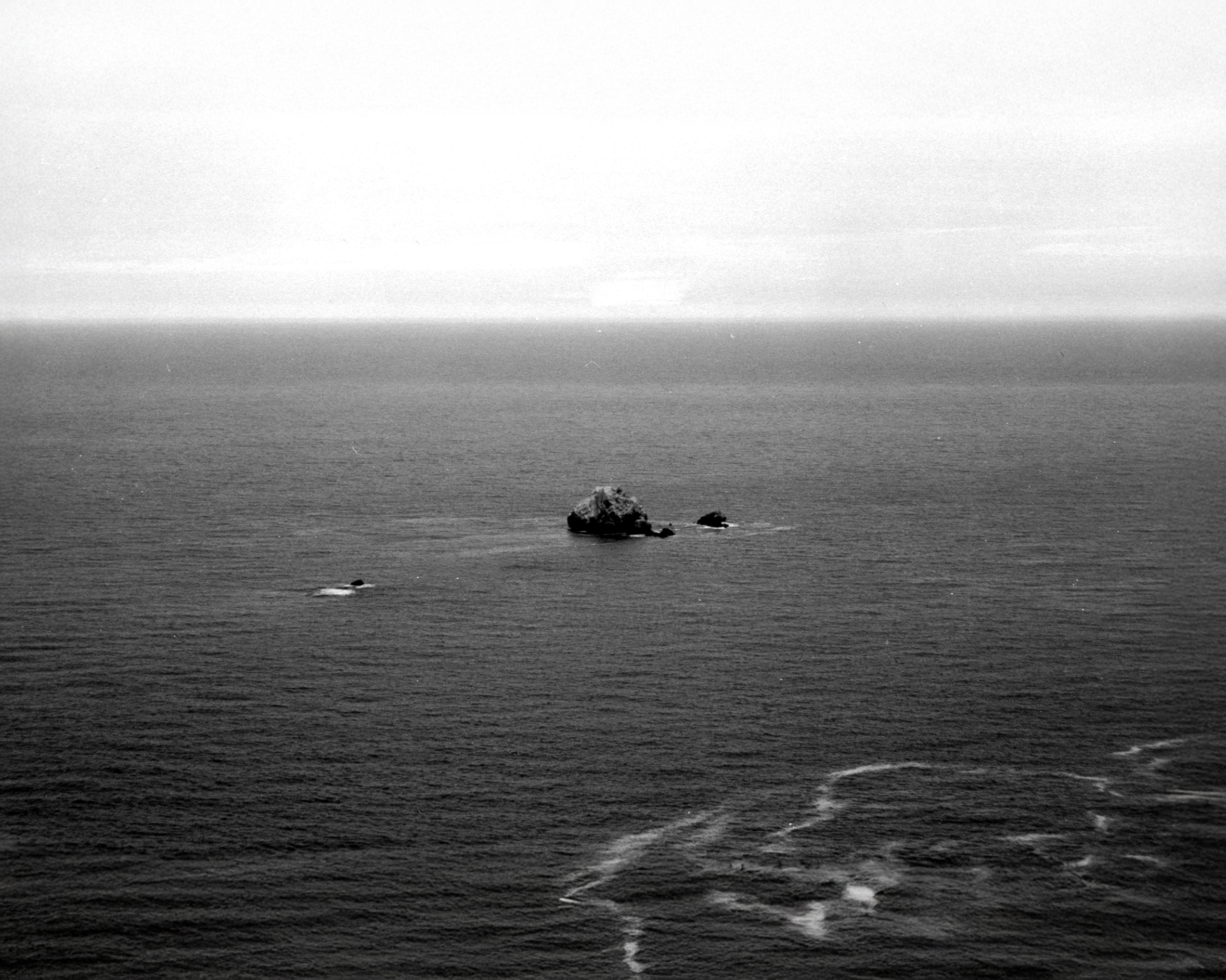 Fotografía en blanco y negro del océano con una gran formación rocosa y un velero a lo lejos.