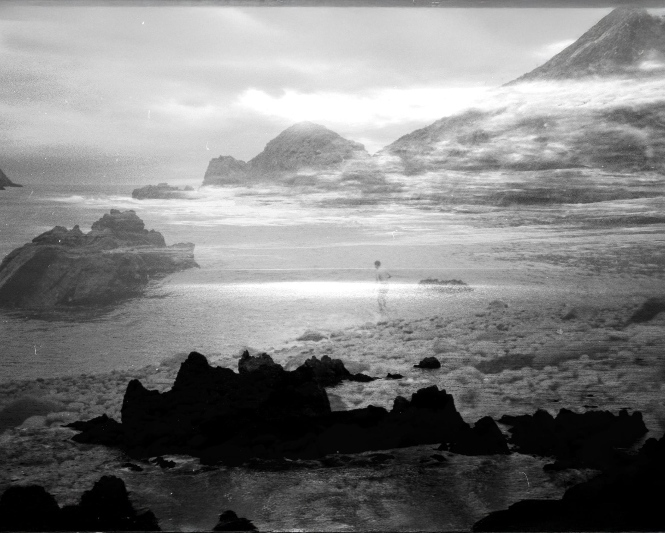 Fotografía en blanco y negro de una costa rocosa con una persona de pie en la playa.
